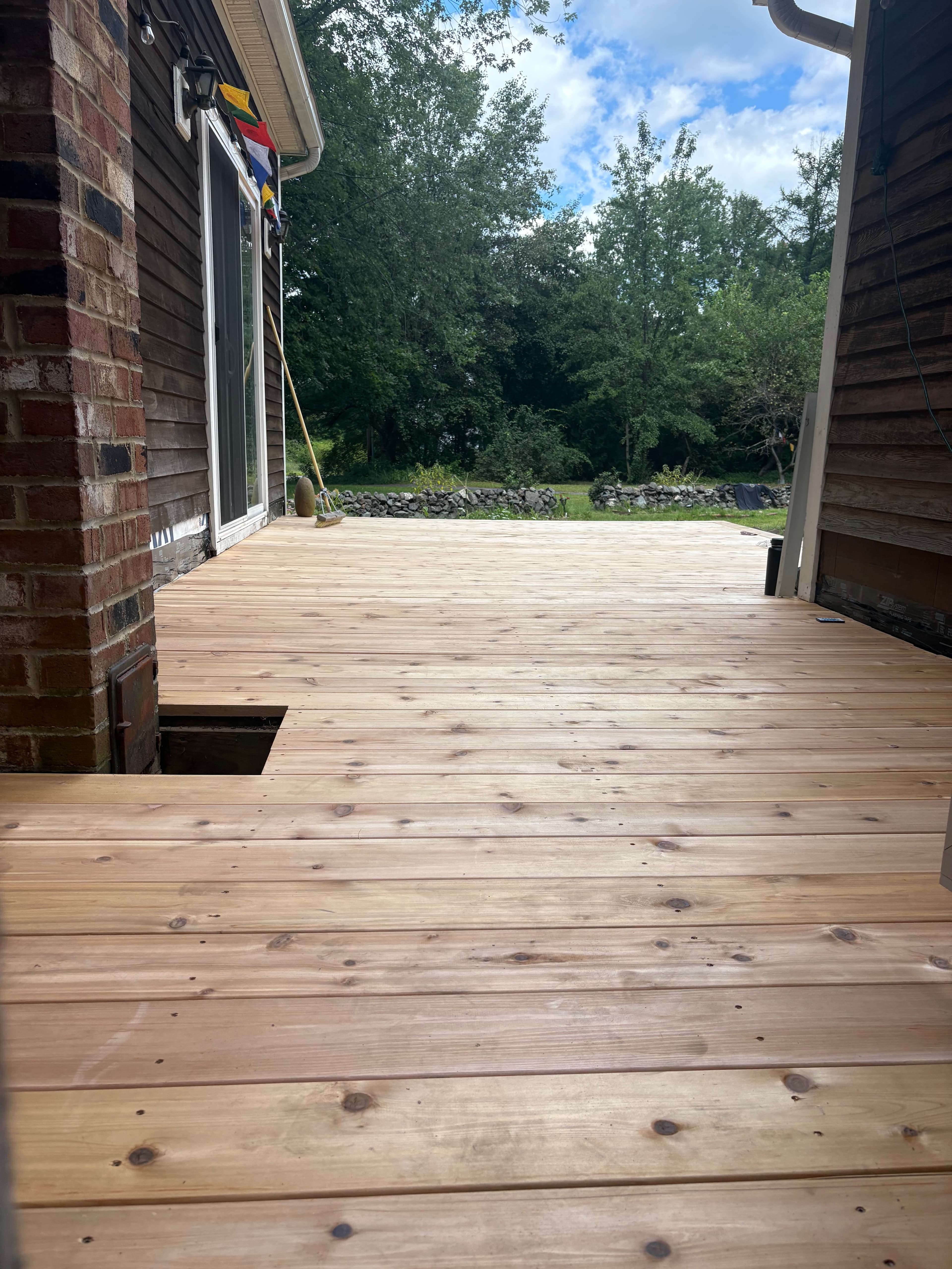 Light wood deck with a square cutout near a brick chimney, overlooking a lush forest.